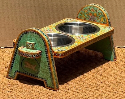 Hand-painted Rajasthani mango wood arch-leg elevated dog bowl stand in sage green with golden ochre floral all-over overlay, orange-red dot-work borders, animal cartouche bracket shelves and two stainless steel bowls